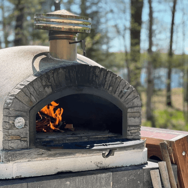 Wood-fired Buena Ventura Preto Premium Pizza Oven with flames inside, set in a forested outdoor area.